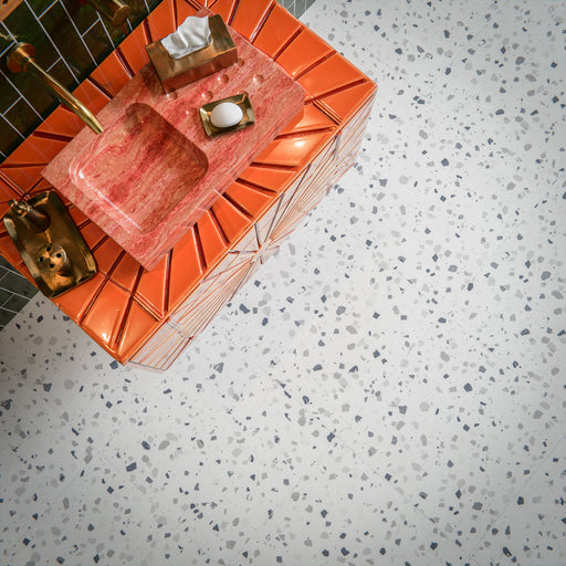 Bathroom interior featuring Veneto Medium White 60 × 60cm terrazzo-effect porcelain floor tiles, showcasing a soft white base with scattered grey stone fragments, paired with Malaga Tourmaline Green gloss brick wall tiles, a statement coral vanity unit, and brushed brass fittings for a bold yet balanced contemporary space.