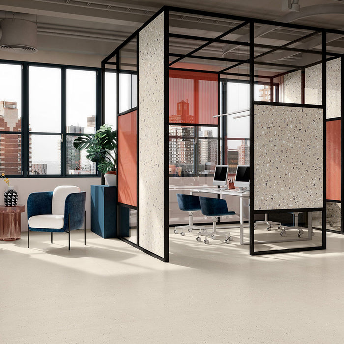 Modern commercial interior featuring Veneto Micro Sand concrete effect porcelain floor tiles, creating a bright, minimalist workspace with black-framed glass partitions, terrazzo effect feature panels, blue upholstered seating and warm contemporary accents.