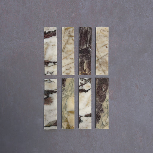 Group of Fitzrovia Viola 5x20cm honed natural marble brick tiles arranged in a neat layout, showcasing a white base with varied burgundy, gold, and grey veining, and a smooth, refined finish.