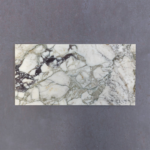 Fitzrovia Viola 30.5x61cm natural marble tiles with a honed finish, featuring a white base with striking green and burgundy veining, shown as a single tile sample laid horizontally against a dark textured backdrop.