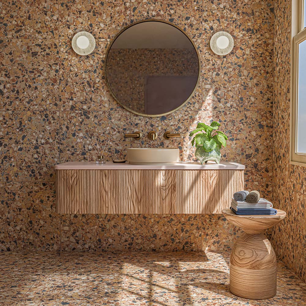 Bathroom featuring Vito Umbar 60cm x 60cm terrazzo style tiles across the walls and floor, showcasing a rich warm-toned terrazzo design with multicoloured stone details, paired with a textured wood vanity, brass fixtures and natural accessories.