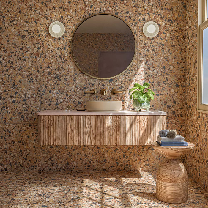 Bathroom featuring Vito Umbar 60cm x 60cm terrazzo style tiles across the walls and floor, showcasing a rich warm-toned terrazzo design with multicoloured stone details, paired with a textured wood vanity, brass fixtures and natural accessories.