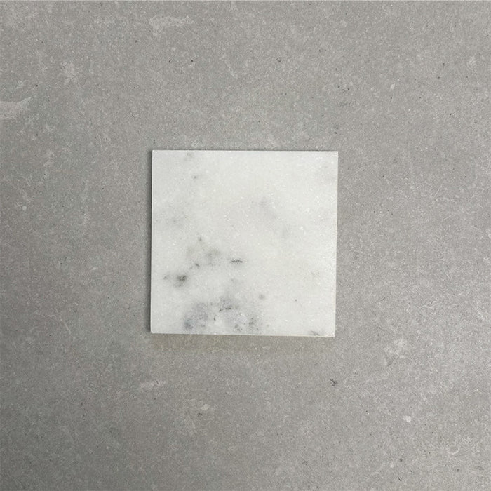 Single Fitzrovia White 15.2x15.2cm honed natural marble tile displayed on a neutral grey surface, showcasing soft white tones, subtle grey veining, and a smooth, refined finish.