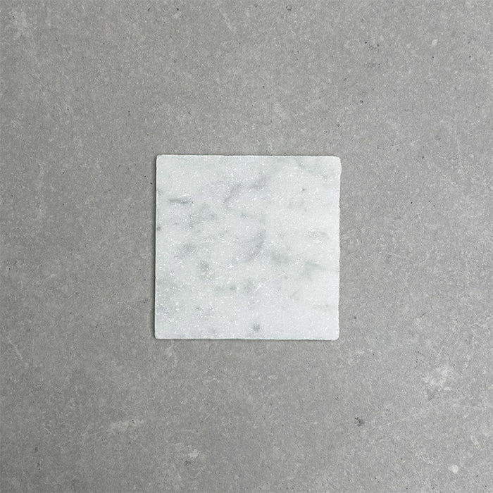 Single Fitzrovia White 15.2x15.2cm tumbled natural marble tile displayed on a neutral grey surface, showcasing soft white tones, subtle grey veining, and a gently aged, textured finish.