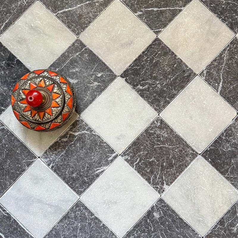 Flat lay of Fitzrovia White 15.2x15.2cm tumbled natural marble tiles arranged in a classic chequerboard pattern with dark marble, showcasing soft tonal variation, aged texture, and a decorative ceramic dish accent.