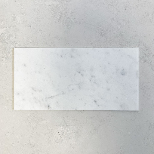 Fitzrovia White 30.5x61cm natural marble tiles with a honed finish, featuring soft white tones with delicate grey veining, shown as a single tile sample laid horizontally against a light textured backdrop.