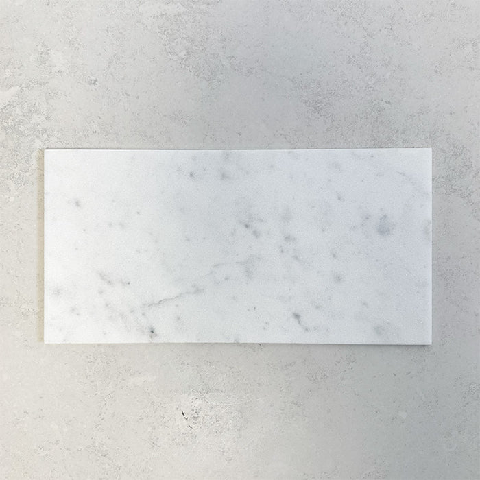 Fitzrovia White 30.5x61cm natural marble tiles with a honed finish, featuring soft white tones with delicate grey veining, shown as a single tile sample laid horizontally against a light textured backdrop.