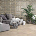 Living room featuring Ziza Emerald Green Moroccan style decorative porcelain wall tiles, showcasing an intricate geometric floral pattern in rich emerald green, charcoal, beige and cream tones with an aged finish, paired with warm stone effect floor tiles, soft grey furnishings, natural textures and lush greenery for a relaxed Moroccan-inspired interior.